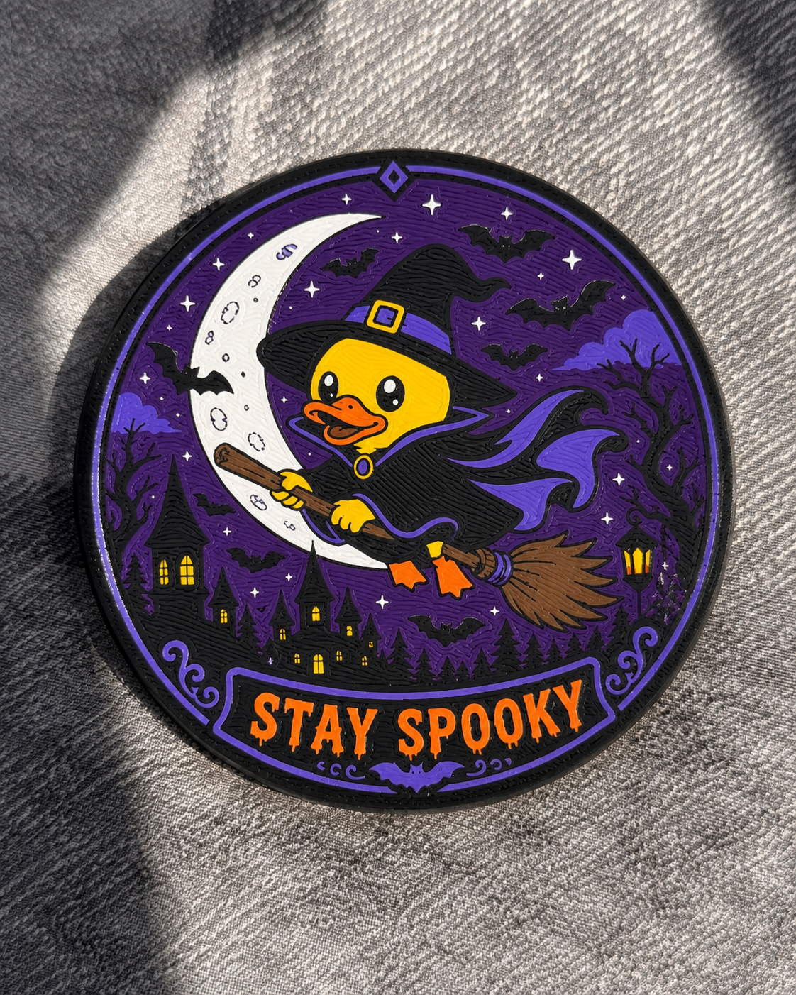 A circular coaster featuring a cartoon yellow duck wearing a witch hat and cape, riding a broomstick under a crescent moon with haunted houses and bats on a purple night sky. The patch reads 'STAY SPOOKY' in orange text and is photographed on a gray textured fabric.